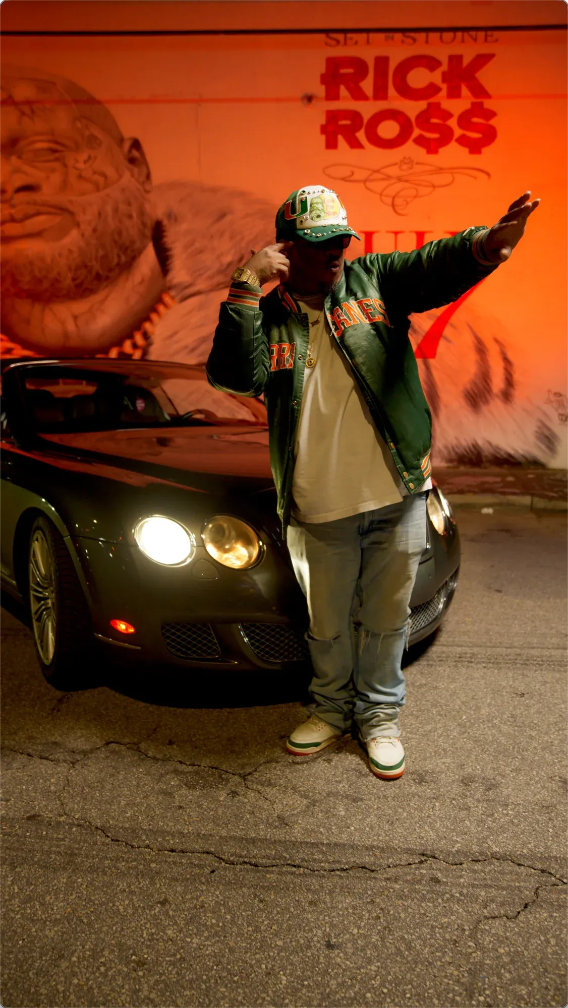 DMONEY3M standing full length in front of a car and the red-lit Rick Ross mural and lettering.